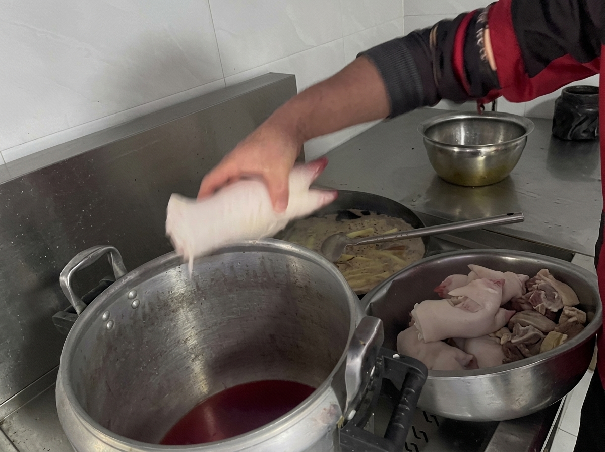 Une main dépose délicatement des morceaux de pieds de porc crus dans une grande marmite remplie de liquide de braisage sombre.