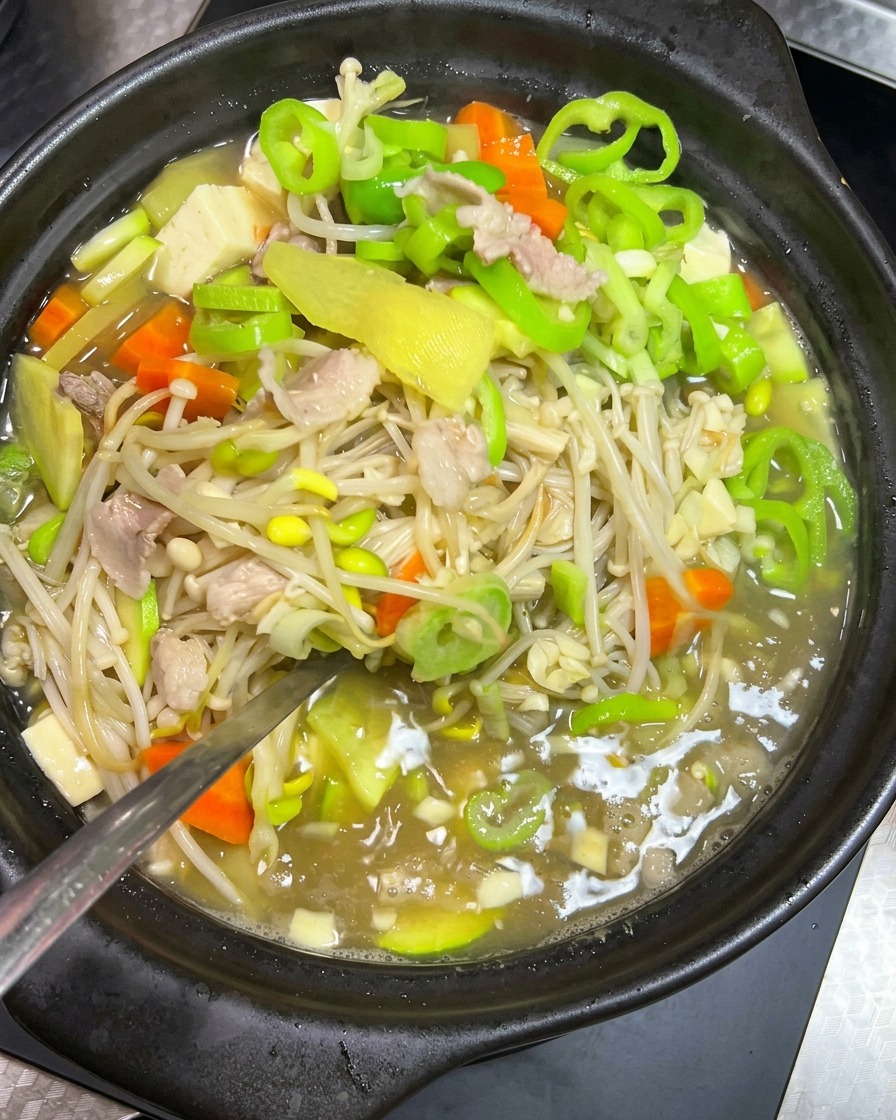 Une cuillère métallique mélangeant doucement un ragoût de pousses de soja, porc et légumes verts dans un pot en terre noir.
