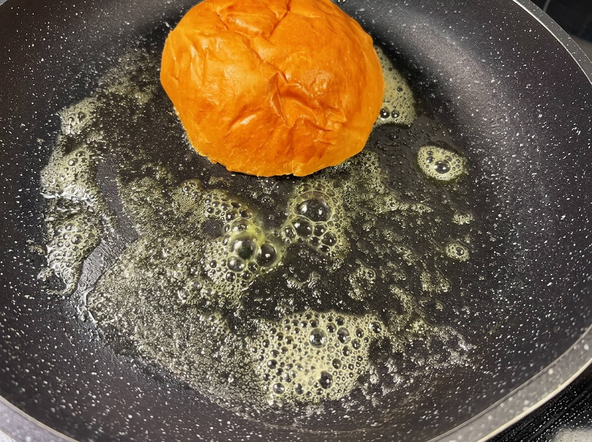 Un pain à burger en train de dorer dans une poêle avec du beurre fondu.