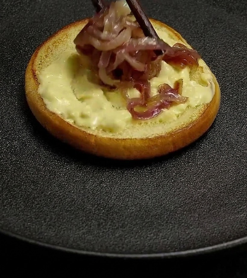 Des oignons rouges cuits placés avec des baguettes sur un pain tartiné de sauce burger crémeuse.