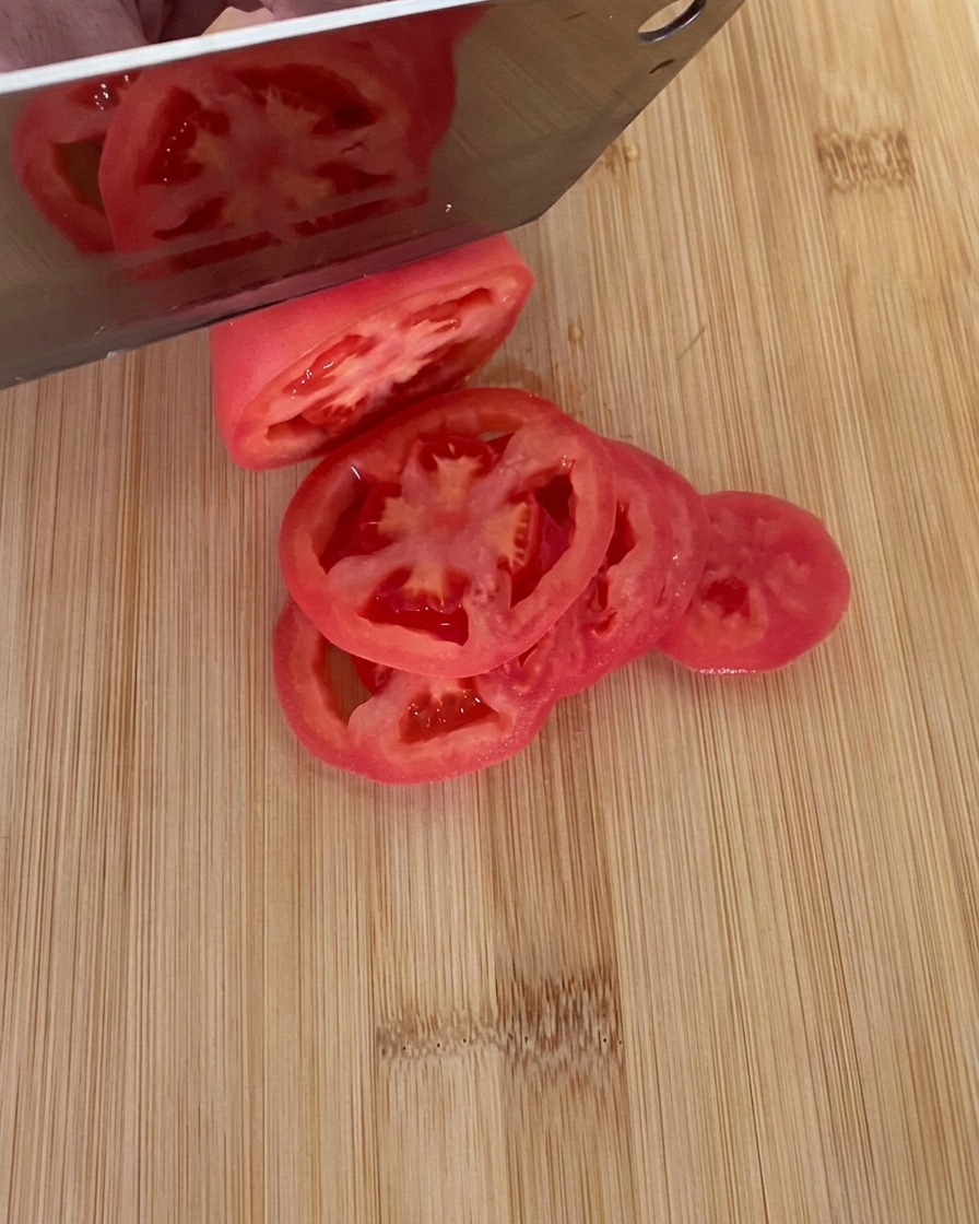 Un couteau découpant une tomate rouge bien mûre en rondelles épaisses sur une planche à découper en bois.