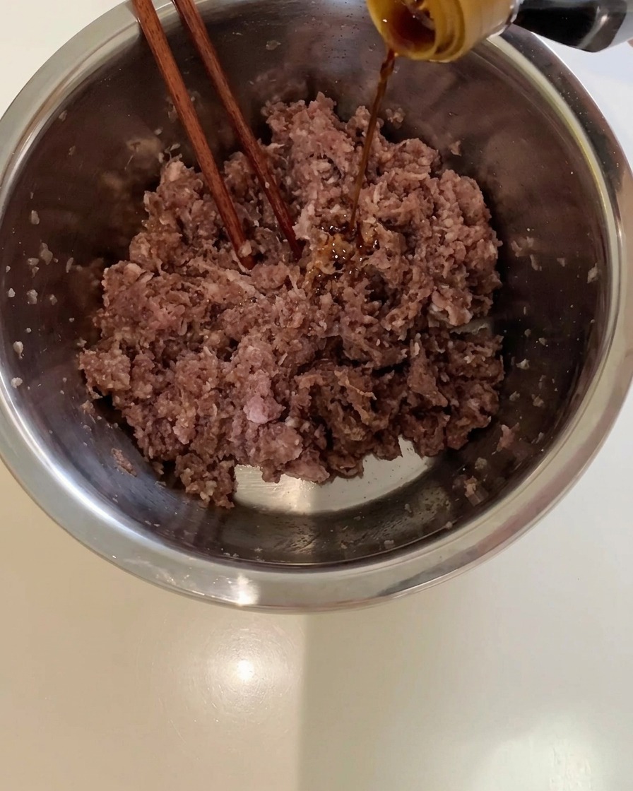 De la sauce soja versée depuis une bouteille dans un bol en métal rempli de viande de bœuf hachée.
