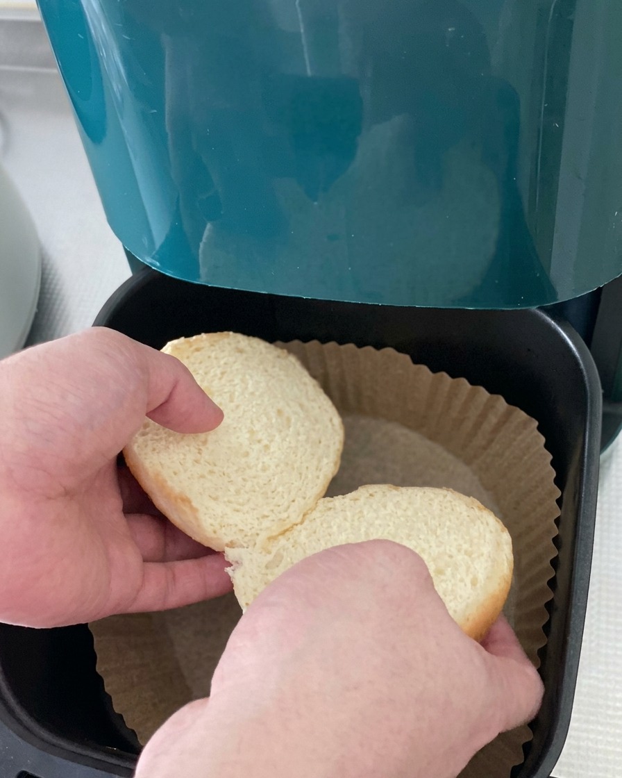 Les deux moitiés dun pain à burger blanc déposées dans le panier dune friteuse à air tapissé de papier cuisson.
