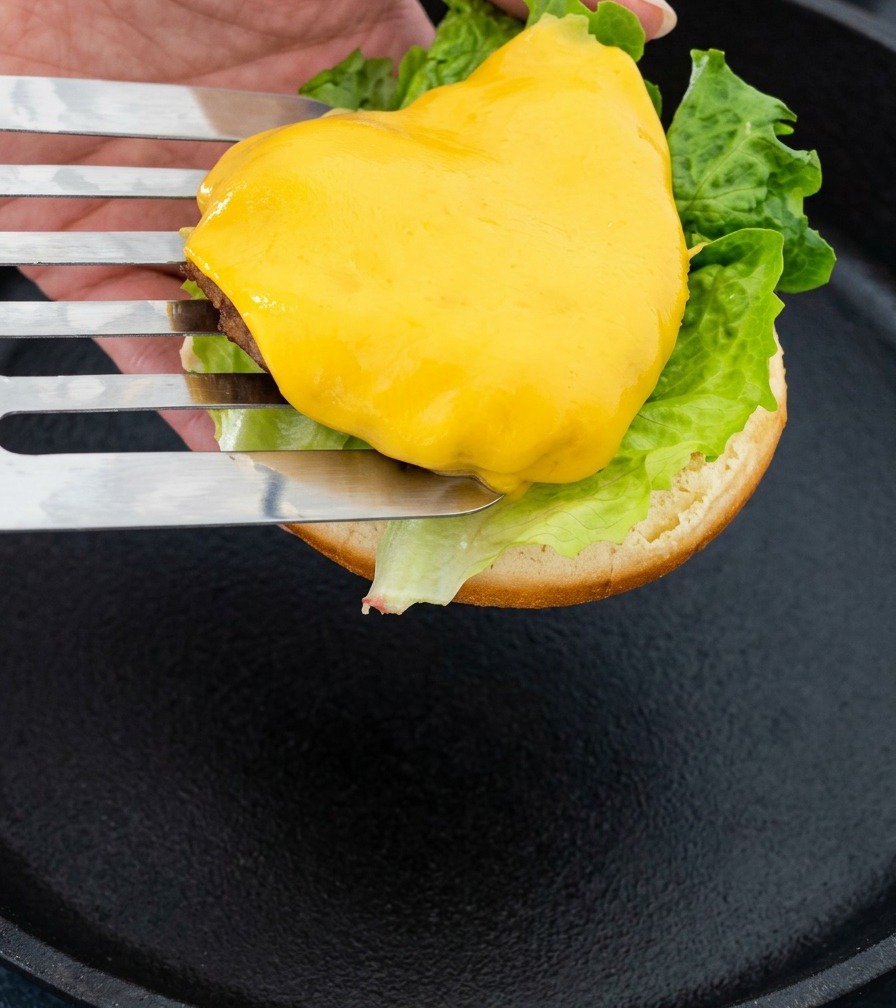 Une spatule en métal soulevant un steak surmonté de fromage fondu pour le déposer sur le pain à burger.