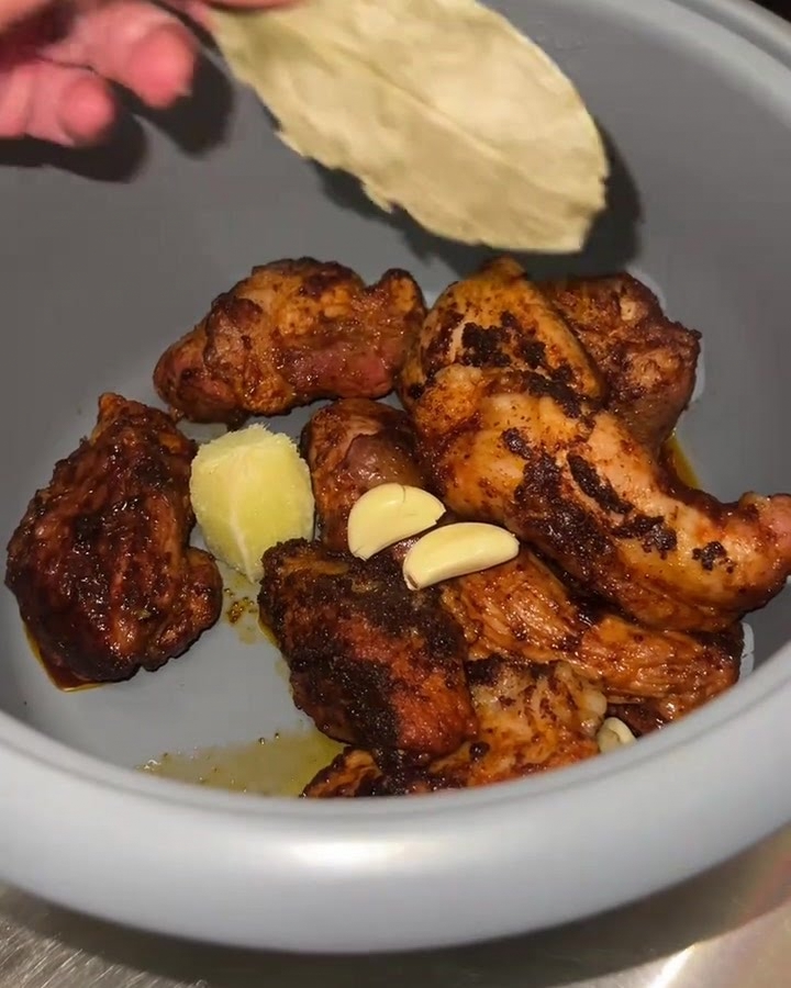 Gros plan sur une cuve de cuiseur à riz contenant des cubes de porc dorés, auxquels on ajoute de lail frais, de la pulpe de citron vert et une feuille de laurier.