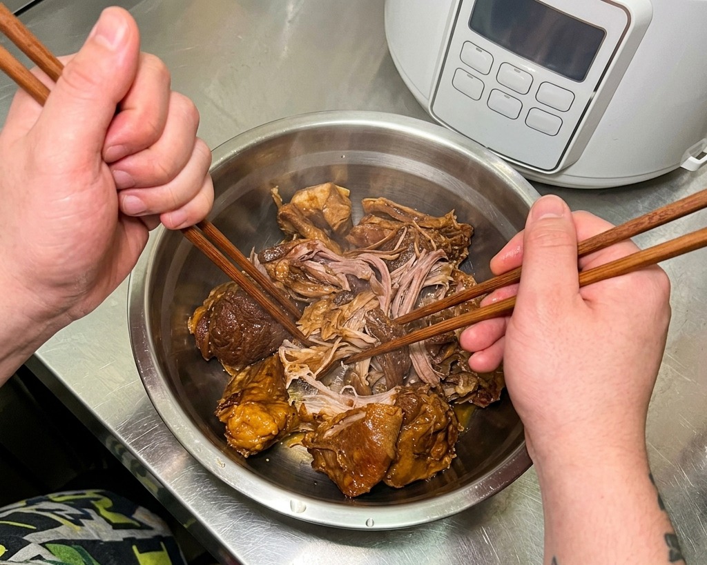 Des mains utilisent des baguettes en bois pour séparer les morceaux de porc braisé très tendre à lintérieur dun bol à mélanger en acier inoxydable.