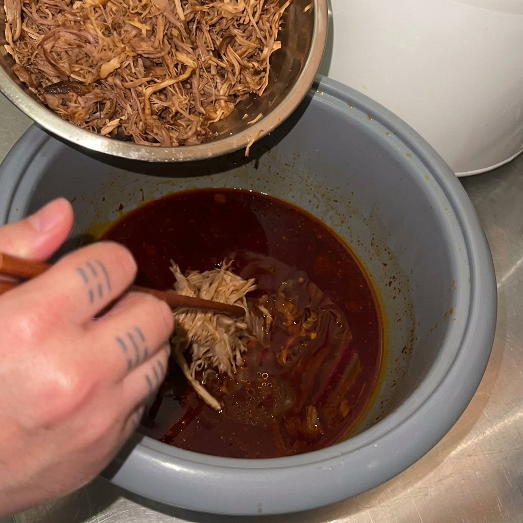 Le porc effiloché est transvasé dun bol en métal vers la cuve du cuiseur à riz contenant la sauce brune de cuisson.