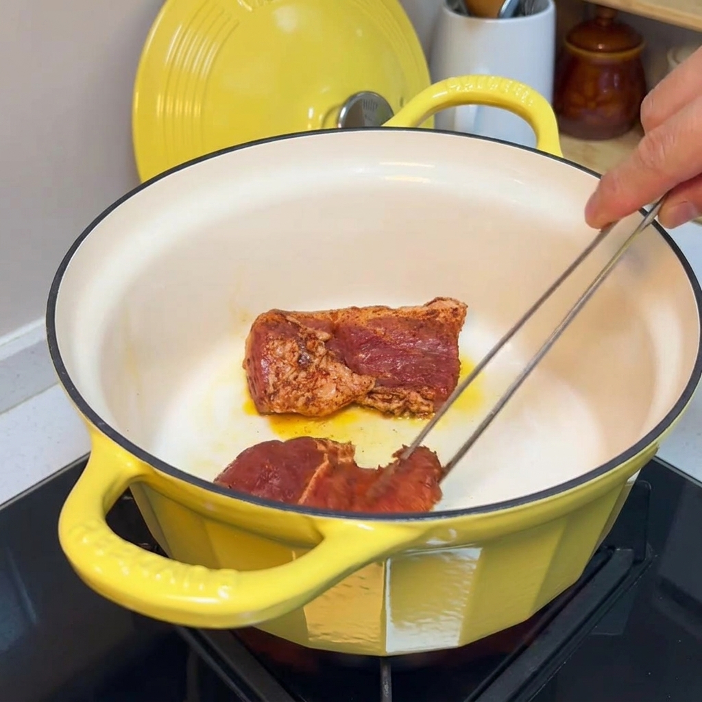 Pince retournant des morceaux de porc assaisonnés dans une cocotte en fonte jaune pour faire dorer les bords.