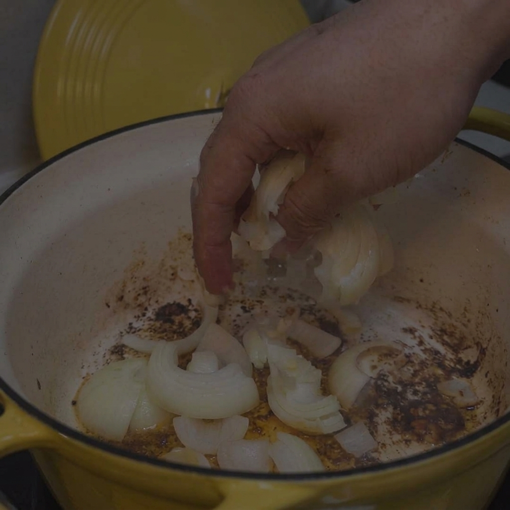 Une main ajoutant des oignons blancs émincés dans une cocotte jaune avec des sucs de cuisson au fond.