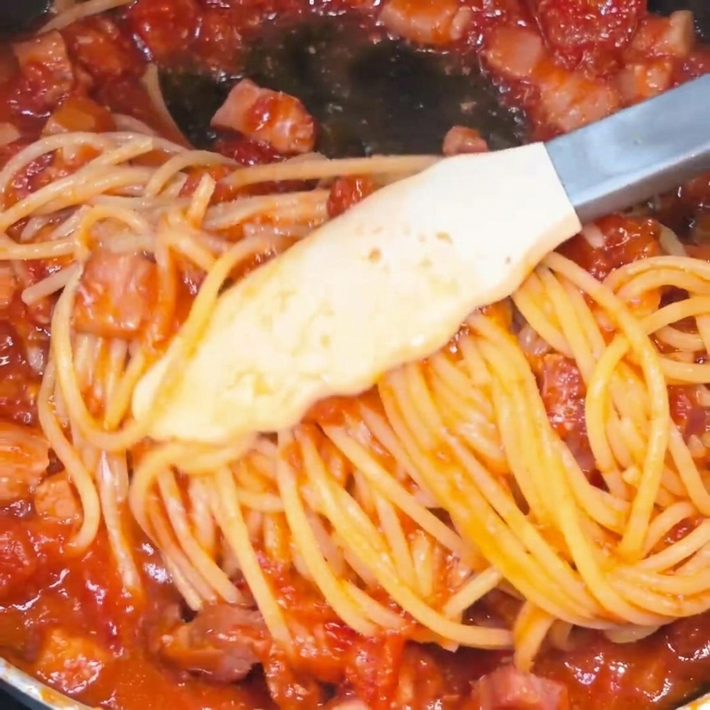 Spaghetti mélangés avec des pinces dans une poêle, parfaitement enrobés dune sauce tomate au bacon rouge vif.
