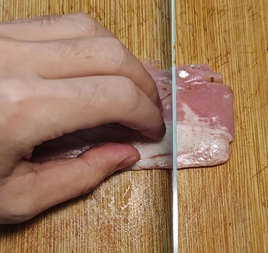 Tranches de bacon coupées en petits morceaux uniformes sur une planche en bois avec un couteau de cuisine.