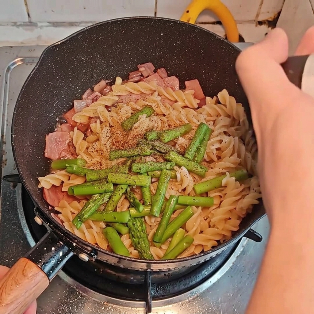 Différents assaisonnements ajoutés sur les pâtes, le bacon et les asperges dans la poêle.