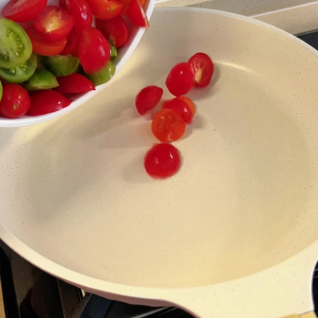 Tomates cerises coupées en deux ajoutées dans une poêle blanche.