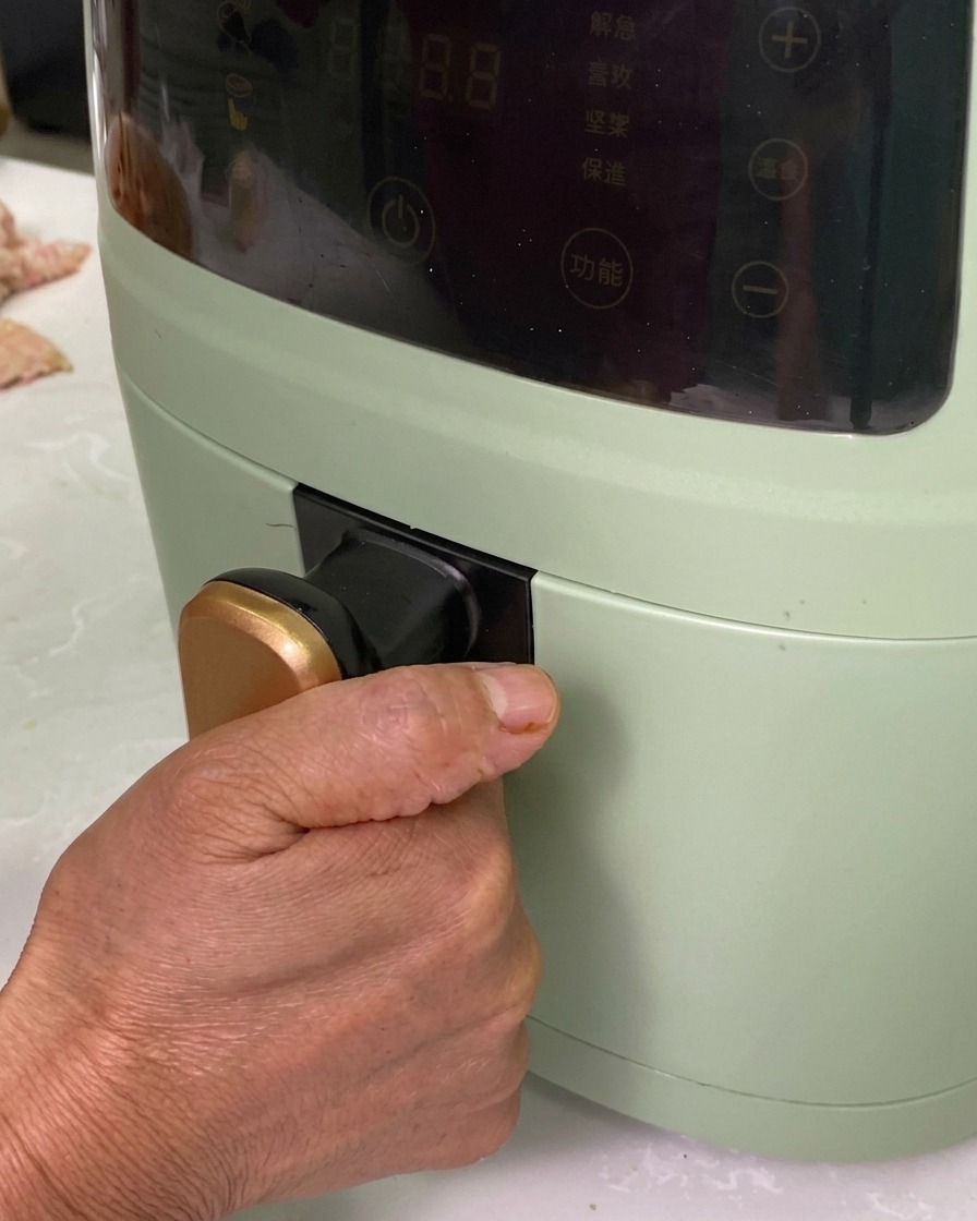 Une main insérant un plat en aluminium garni de roulés de porc assaisonnés dans un air fryer vert.