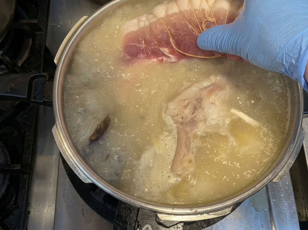 Une main gantée plaçant un morceau déchine de porc ficelée dans une marmite de bouillon bouillant avec des légumes.