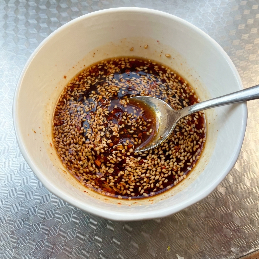 Un petit bol blanc contenant un mélange de sauce épicée sombre parsemé de graines de sésame, en cours de mélange avec une cuillère.