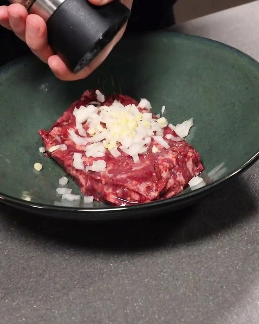 Bœuf haché mélangé avec de lail et de loignon dans un bol, avec ajout de poivre noir.