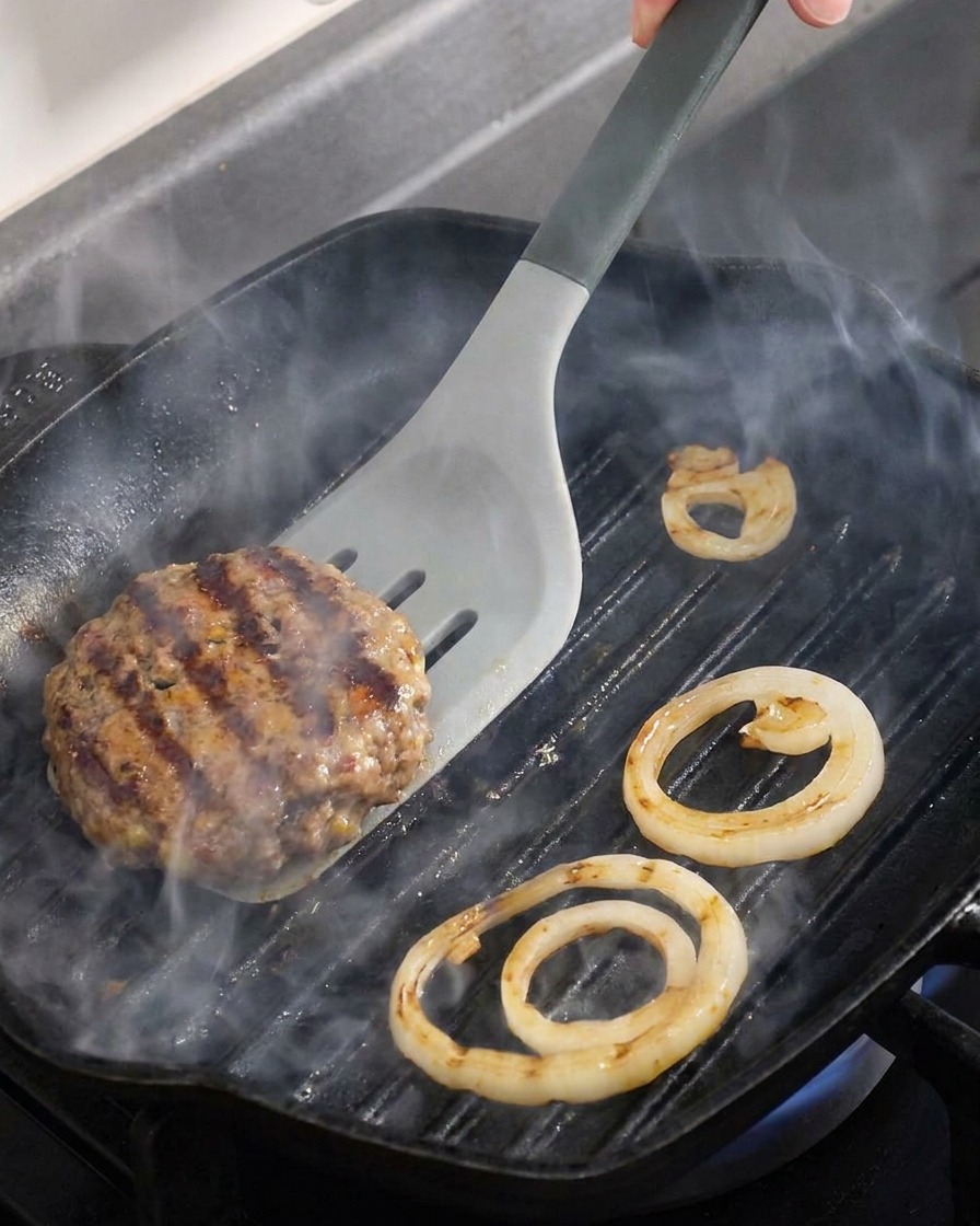 Gros plan dun steak haché et de rondelles doignon retournés dans une poêle à griller bien chaude.