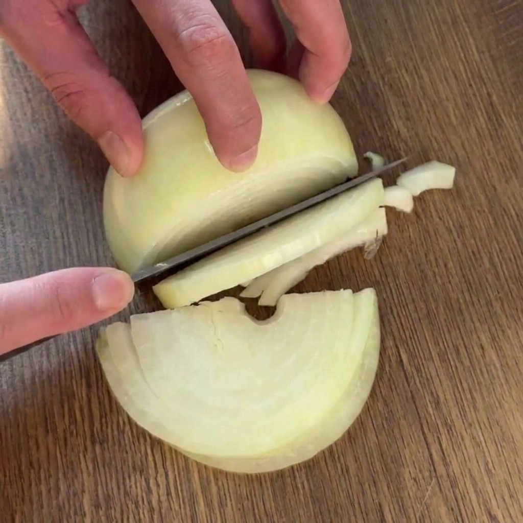 Une personne éminçant un demi-oignon jaune en fines lamelles régulières avec un couteau de chef sur une planche en bois.