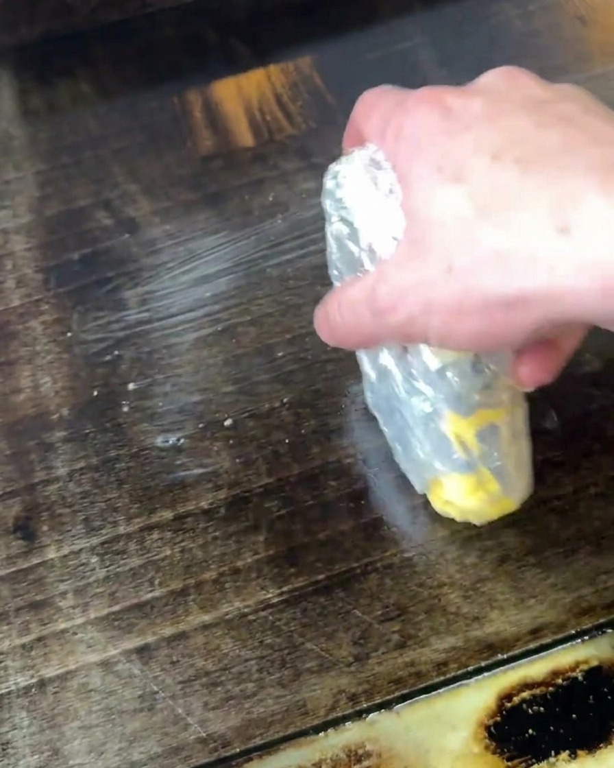Une main tenant une motte de beurre partiellement emballée, la faisant fondre en la frottant directement sur une plancha chaude et sombre.
