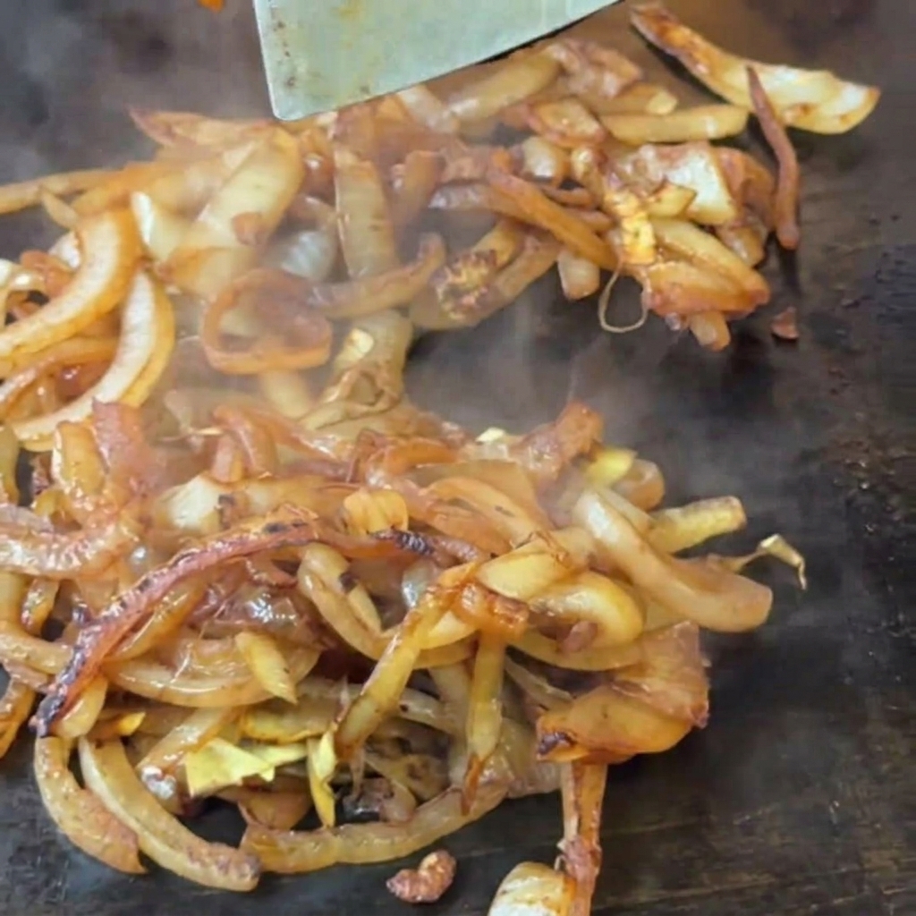 Une spatule en métal remuant des oignons émincés ramollis et légèrement bruns sur une plancha chaude.