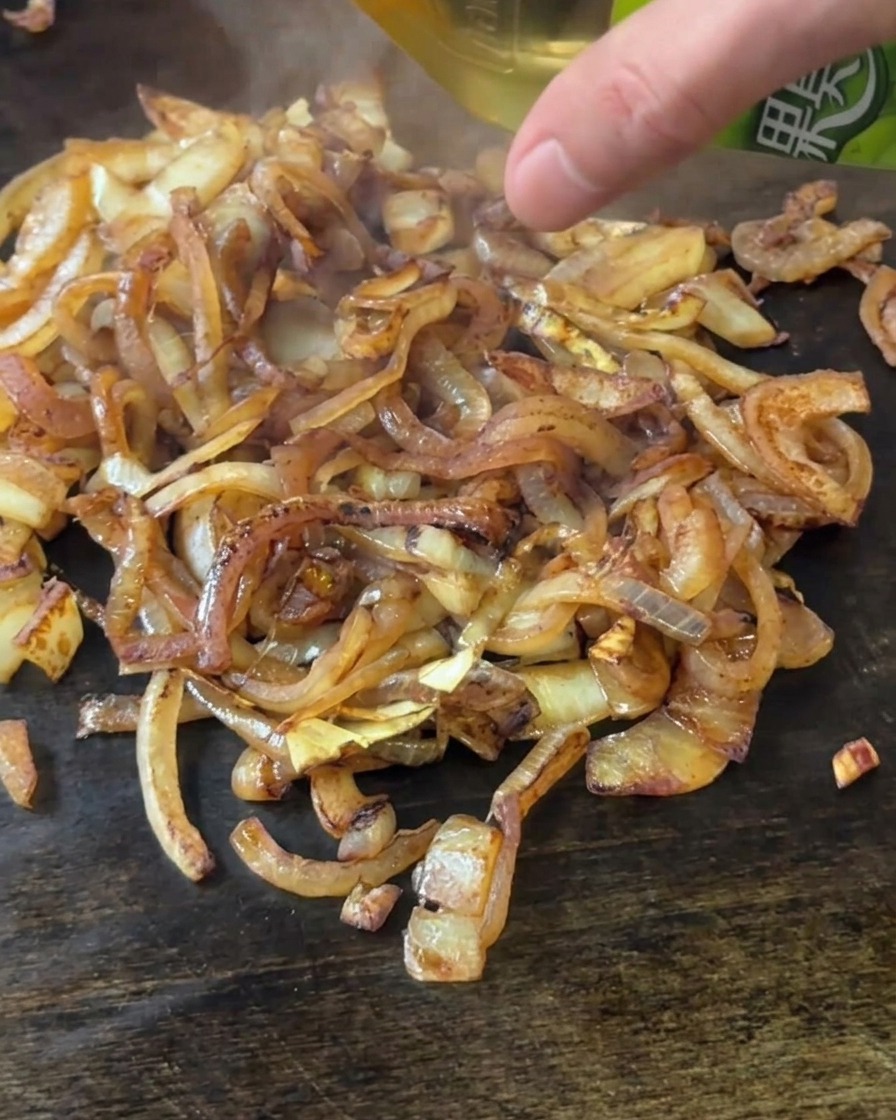 Une main versant du vinaigre de cidre depuis une bouteille verte sur un tas doignons brunis sur une plaque chauffante.