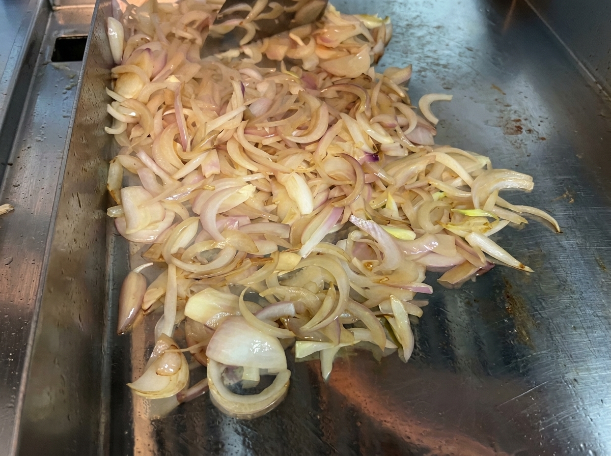 Oignons émincés mélangés continuellement avec une spatule sur une surface de cuisson chaude pour les ramollir et les caraméliser.