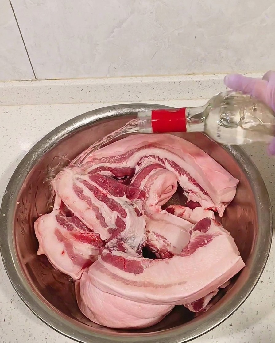 Morceaux de poitrine de porc crue dans un bol en métal, arrosés dalcool blanc.