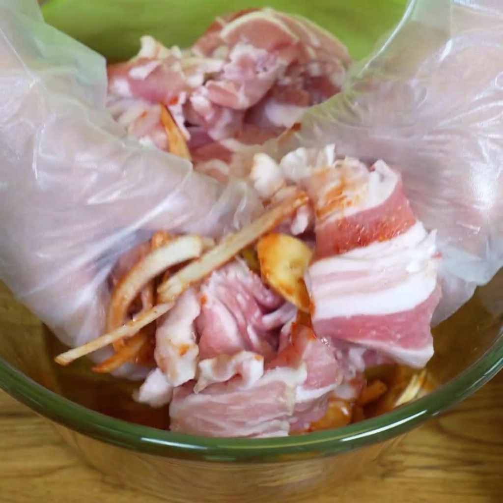 Mains gantées mélangeant le porc, la marinade et les légumes dans un bol en verre.