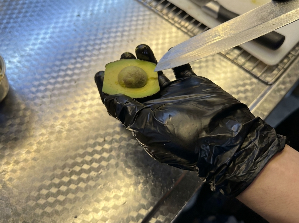 Main avec un gant noir tenant un demi-avocat et retirant le noyau avec un couteau.