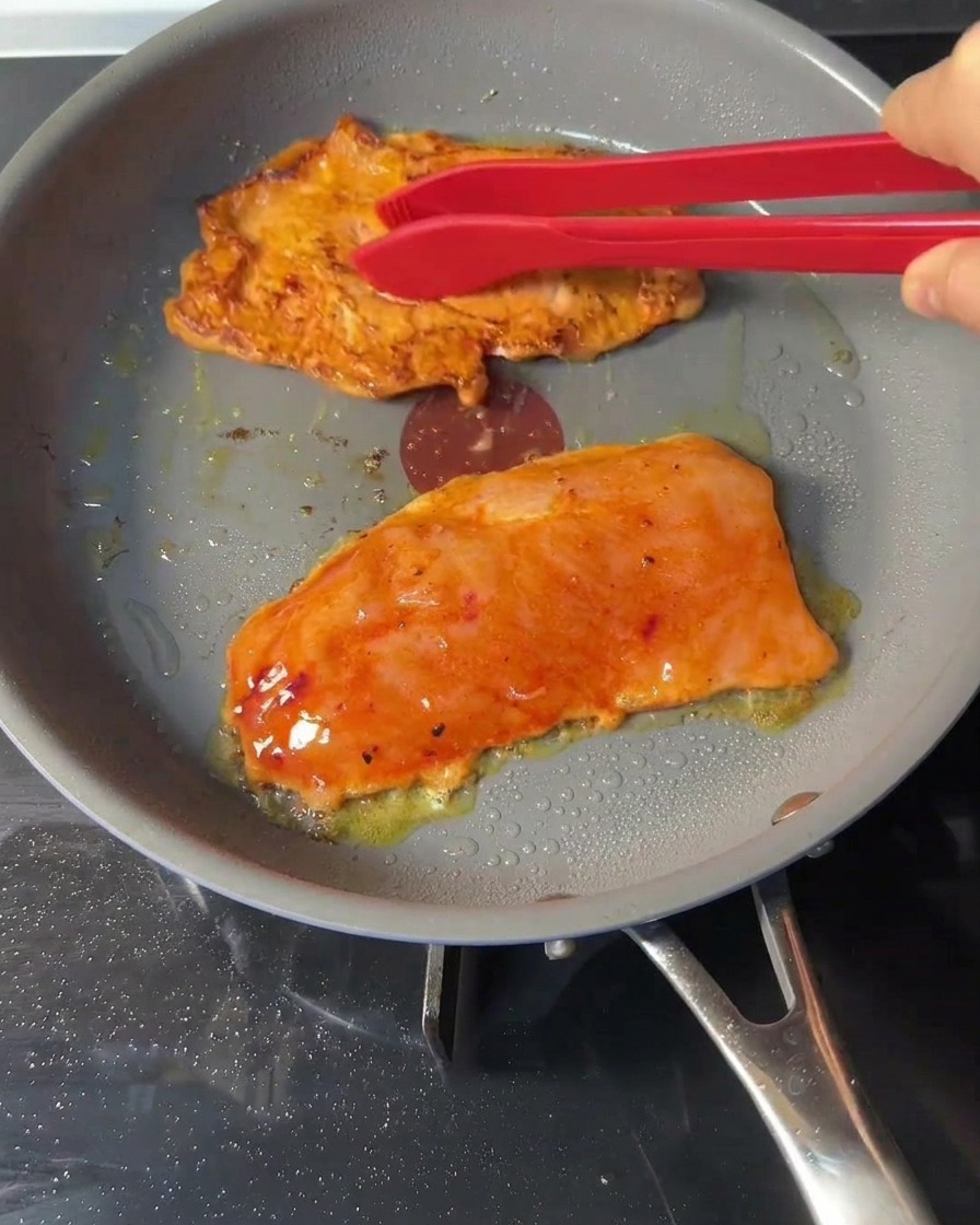 Des pinces rouges retournant des steaks de poulet en cours de cuisson dans une poêle.