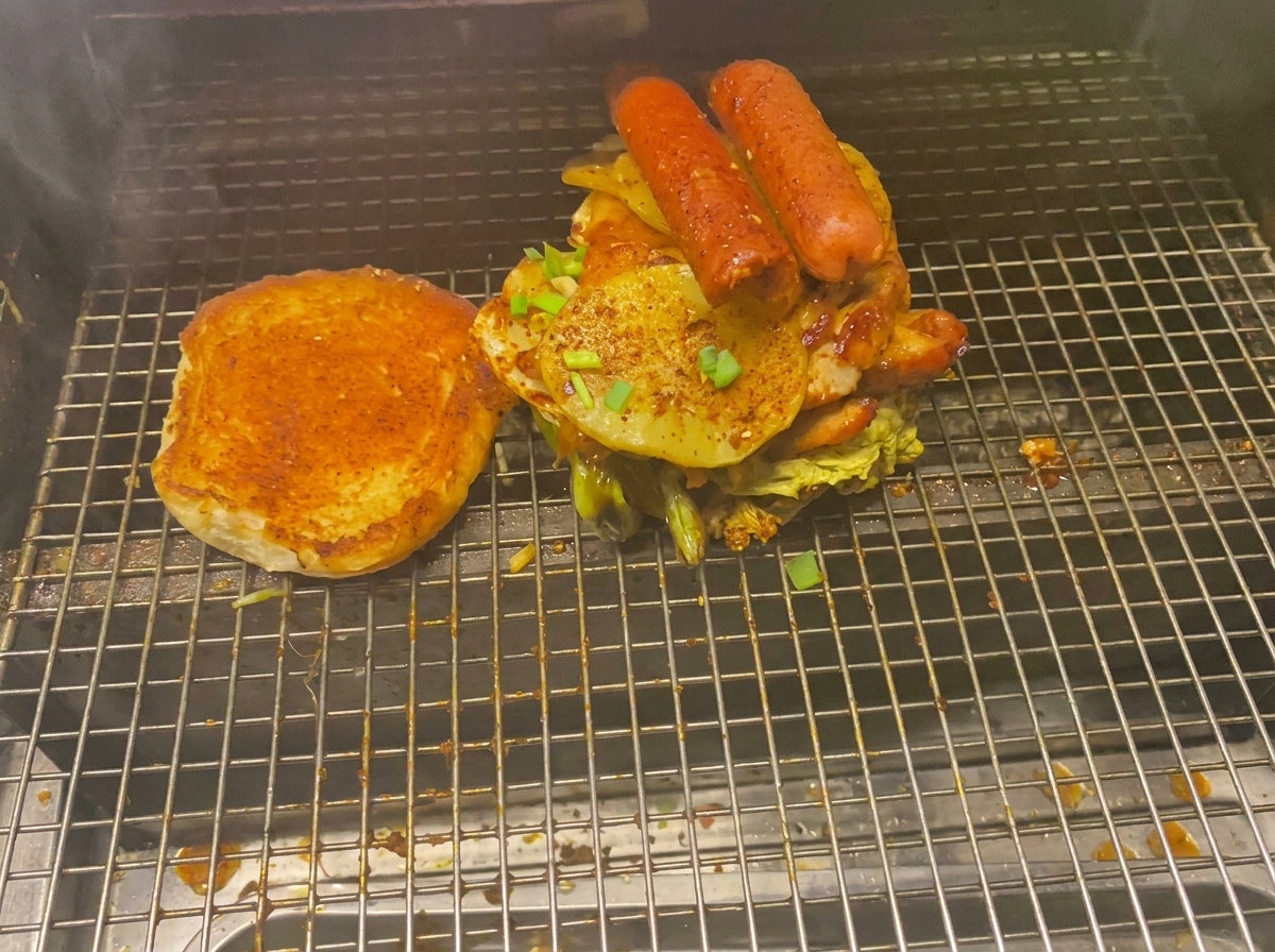 Un burger imposant chargé de viandes, pommes de terre grillées et saucisses à côté du pain sur la grille.