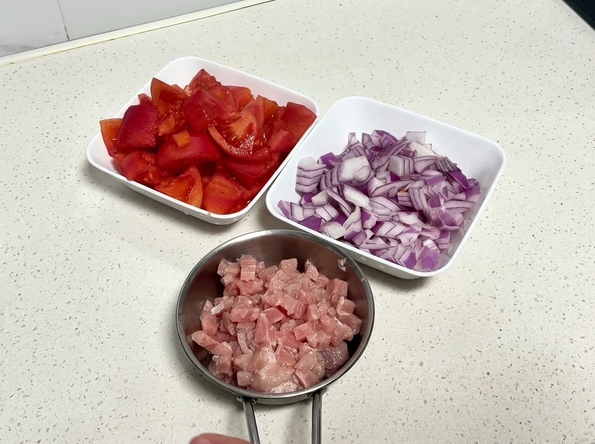 Vue de dessus de trois bols blancs remplis de tomates fraîches coupées, doignon rouge haché et de porc cru en dés sur un plan de travail.