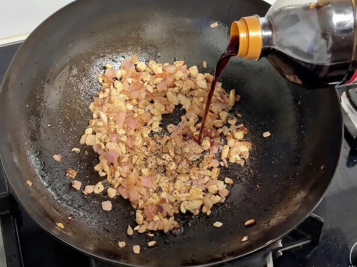 Sauce soja versée dune bouteille dans un wok contenant du porc doré et des oignons, saupoudré de poivre noir.
