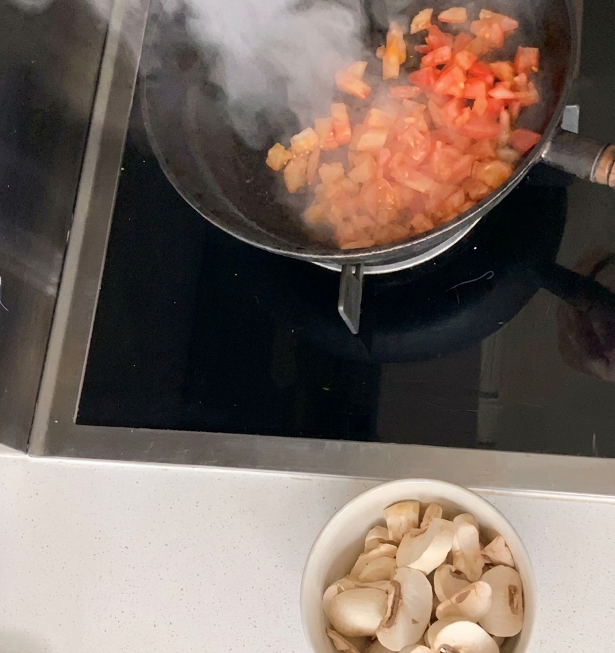 Des dés de tomates sautés dans une poêle noire avec un bol de champignons émincés à côté.