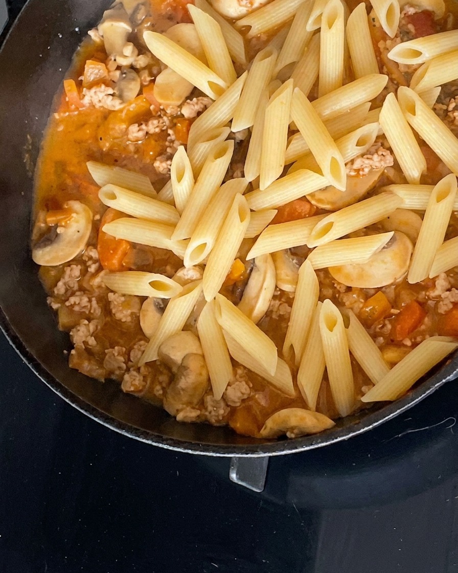 Des penne cuites ajoutées à une savoureuse sauce à la viande hachée, aux champignons et aux tomates dans une poêle noire.