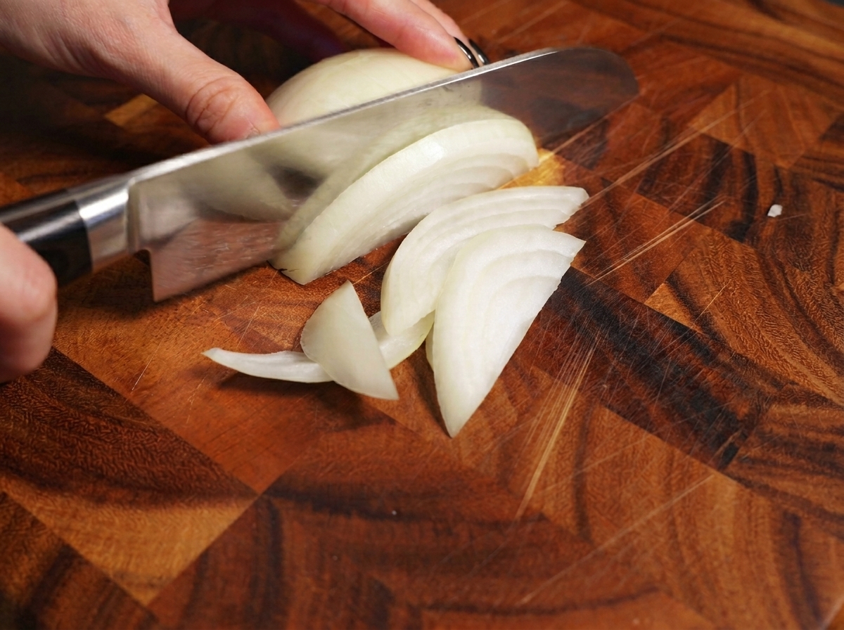 Une main utilisant un couteau de chef pour trancher un oignon blanc sur une planche en bois.