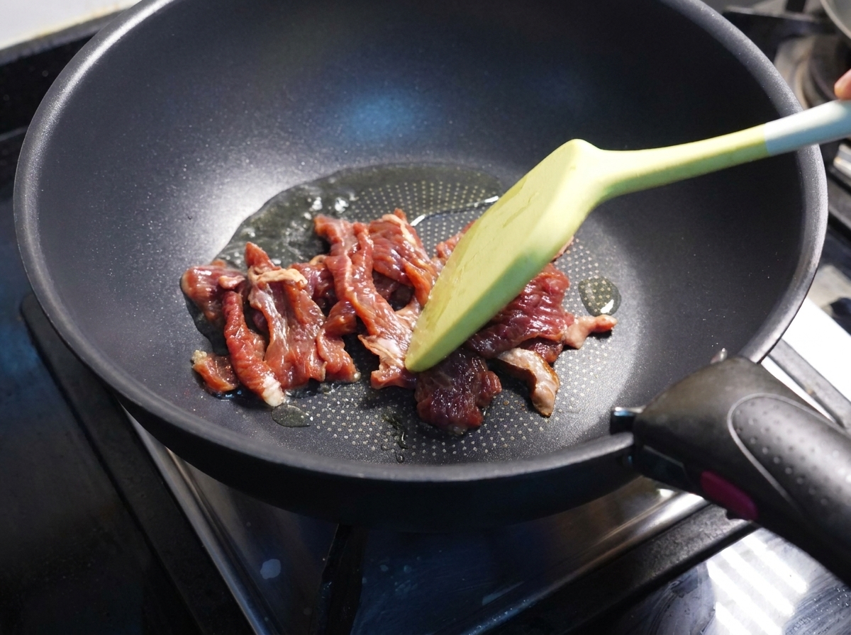 Spatule faisant sauter des tranches de bœuf partiellement cuites dans une poêle sombre.
