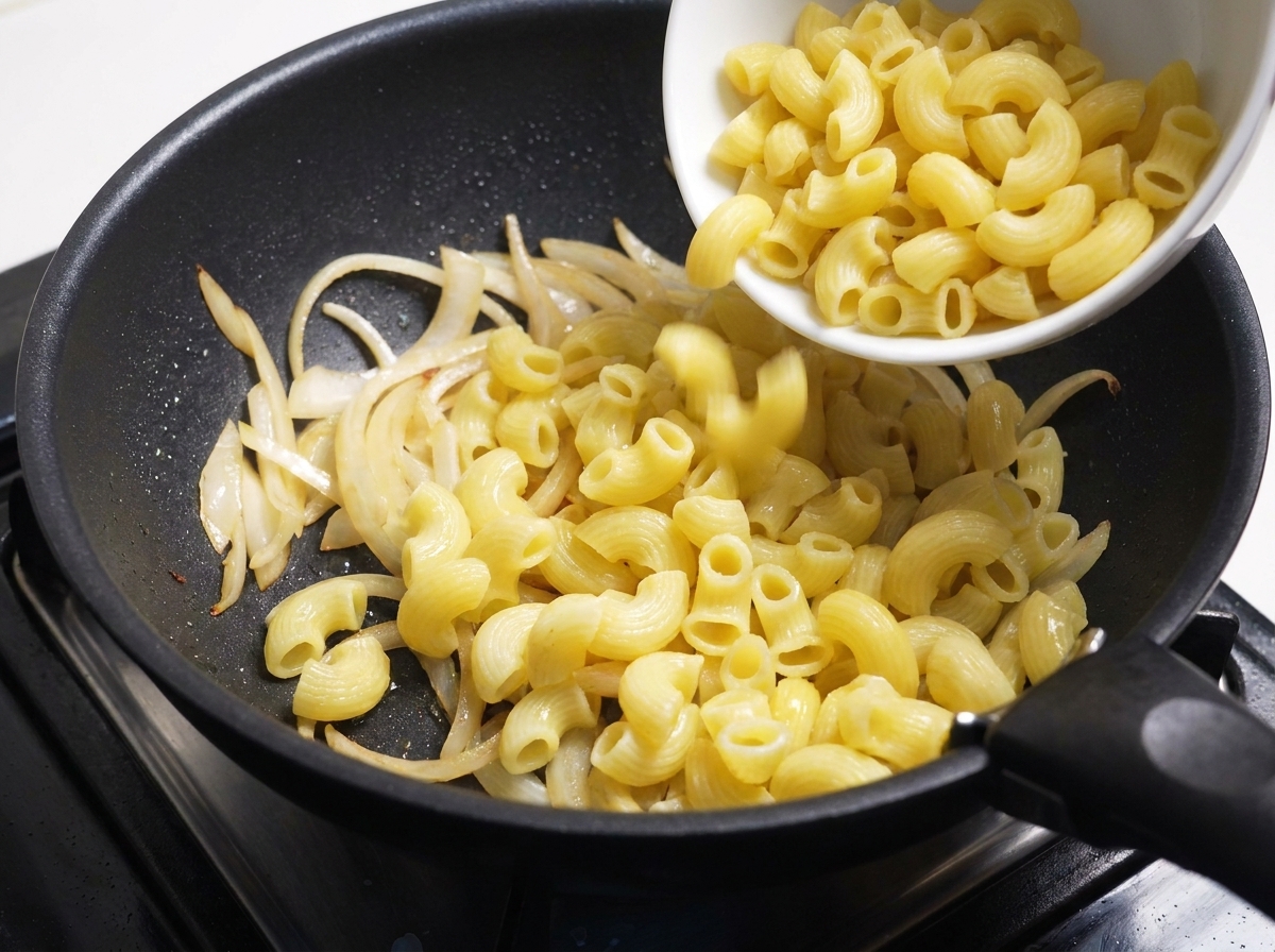 Macaronis cuits versés depuis un bol blanc dans une poêle avec des oignons sautés.
