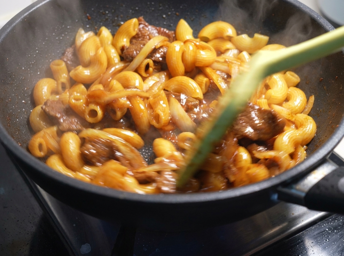 Macaronis, bœuf et oignons mélangés rapidement avec une spatule dans une poêle chaude.