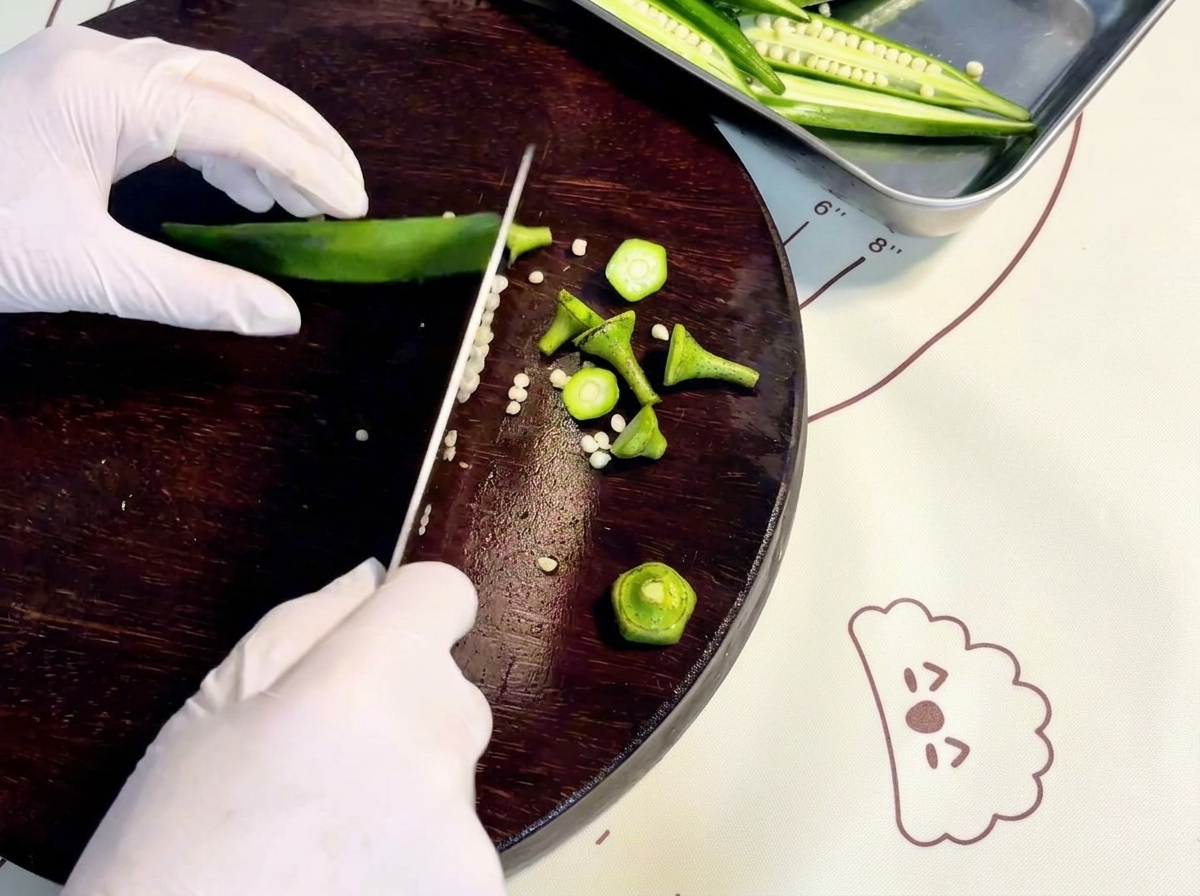 Mains gantées tranchant du gombo vert frais dans le sens de la longueur sur une planche à découper en bois sombre.