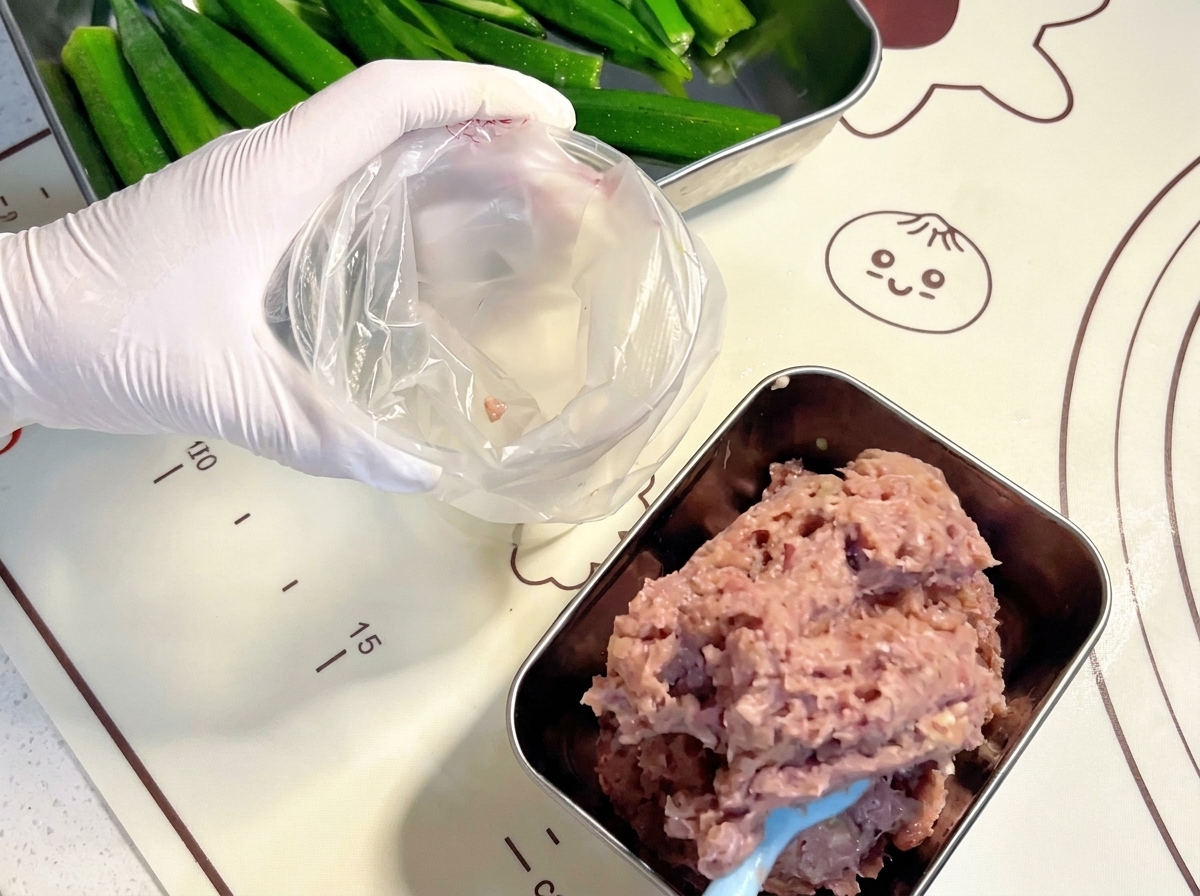 Mains gantées utilisant une spatule bleue pour transférer la pâte de viande crue dans une poche à douille en plastique transparent.
