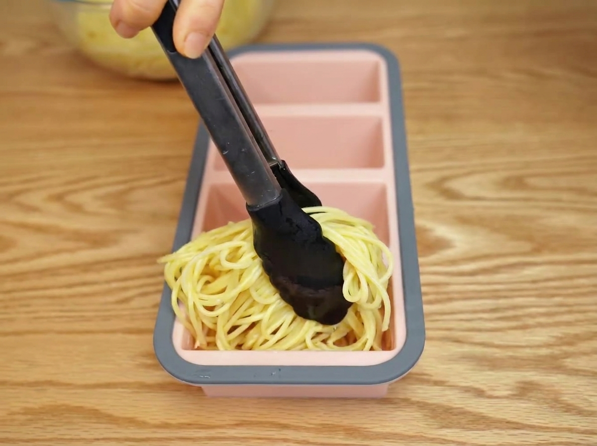 Des spaghettis cuits placés dans un moule en silicone à laide de pinces.