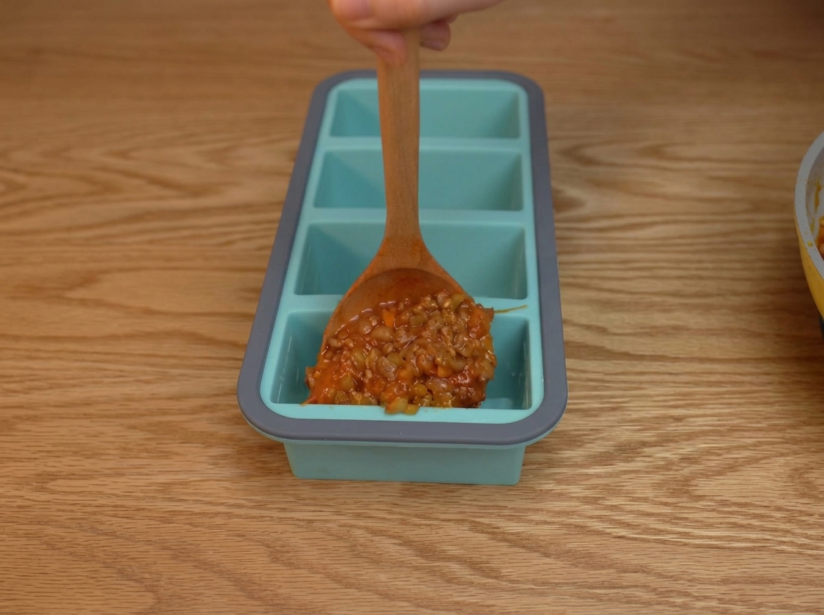 Une épaisse sauce à la viande et à la tomate déposée à la cuillère dans les compartiments individuels dun bac en silicone bleu.