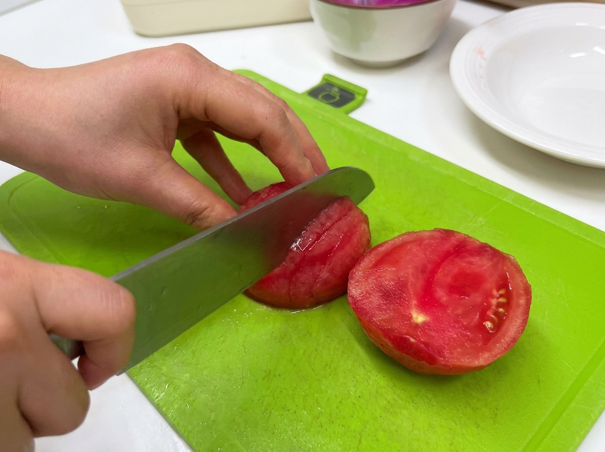 Une personne utilisant un grand couteau de cuisine pour trancher une tomate pelée sur une planche à découper verte.