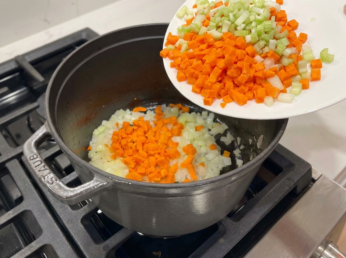 Des carottes et du céleri versés dune assiette blanche dans une marmite contenant des oignons revenus.