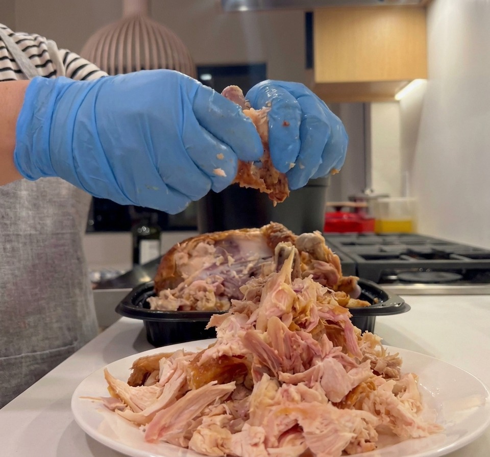 Des mains portant des gants de cuisine bleus effilochant la chair dun poulet rôti sur une assiette blanche.