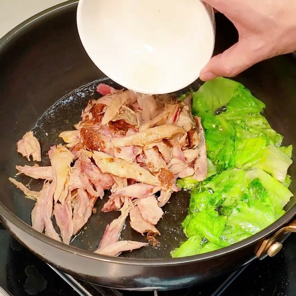 Poulet cuit effiloché versé dun petit bol blanc dans la poêle contenant la laitue sautée.