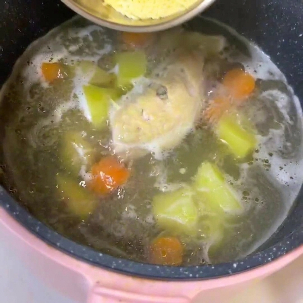 Une casserole de bouillon frémissant contenant du poulet, des dés de pommes de terre et des carottes, dans laquelle on incorpore les nouilles pour bébé.