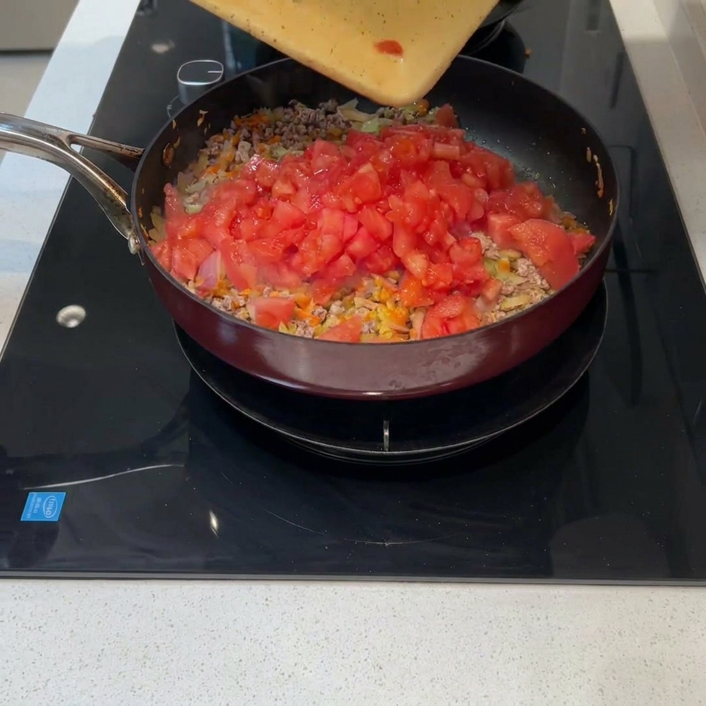Des tomates fraîchement coupées en dés versées depuis une planche à découper dans une poêle contenant la viande et les légumes.